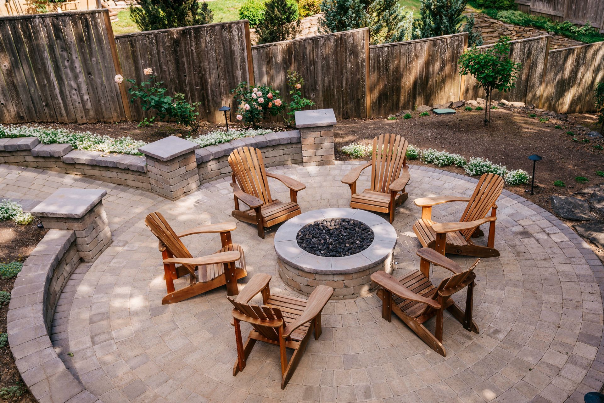 Outdoor patio with Adirondack chairs around a fire pit on a stone surface. Stone retaining walls and wood fence.