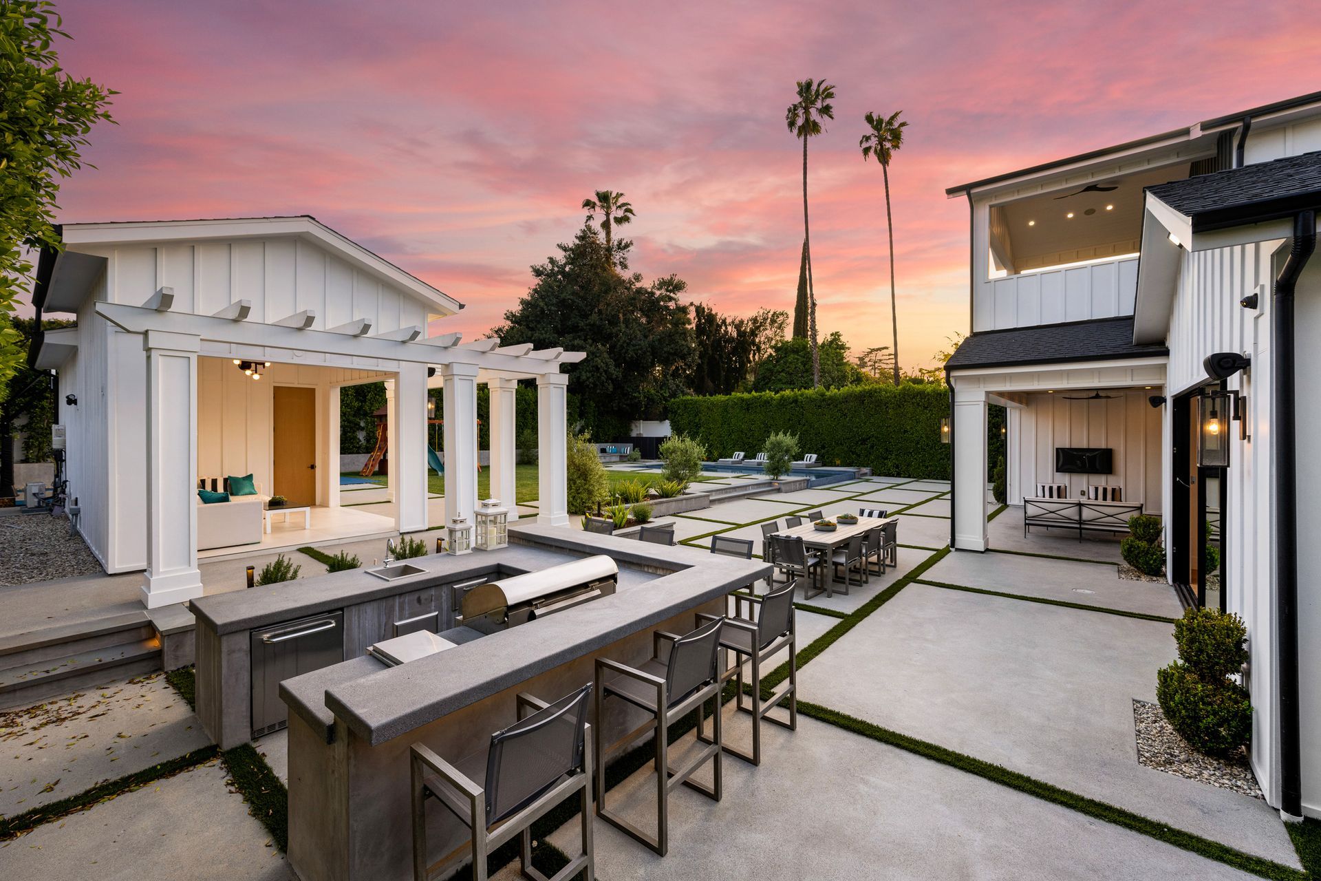 Outdoor kitchen and patio at dusk with a bar, dining table, and building.