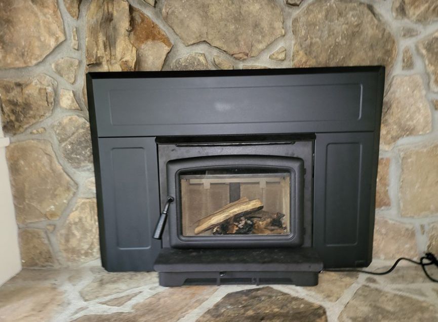 Black fireplace insert in a stone wall, containing logs behind a glass door.