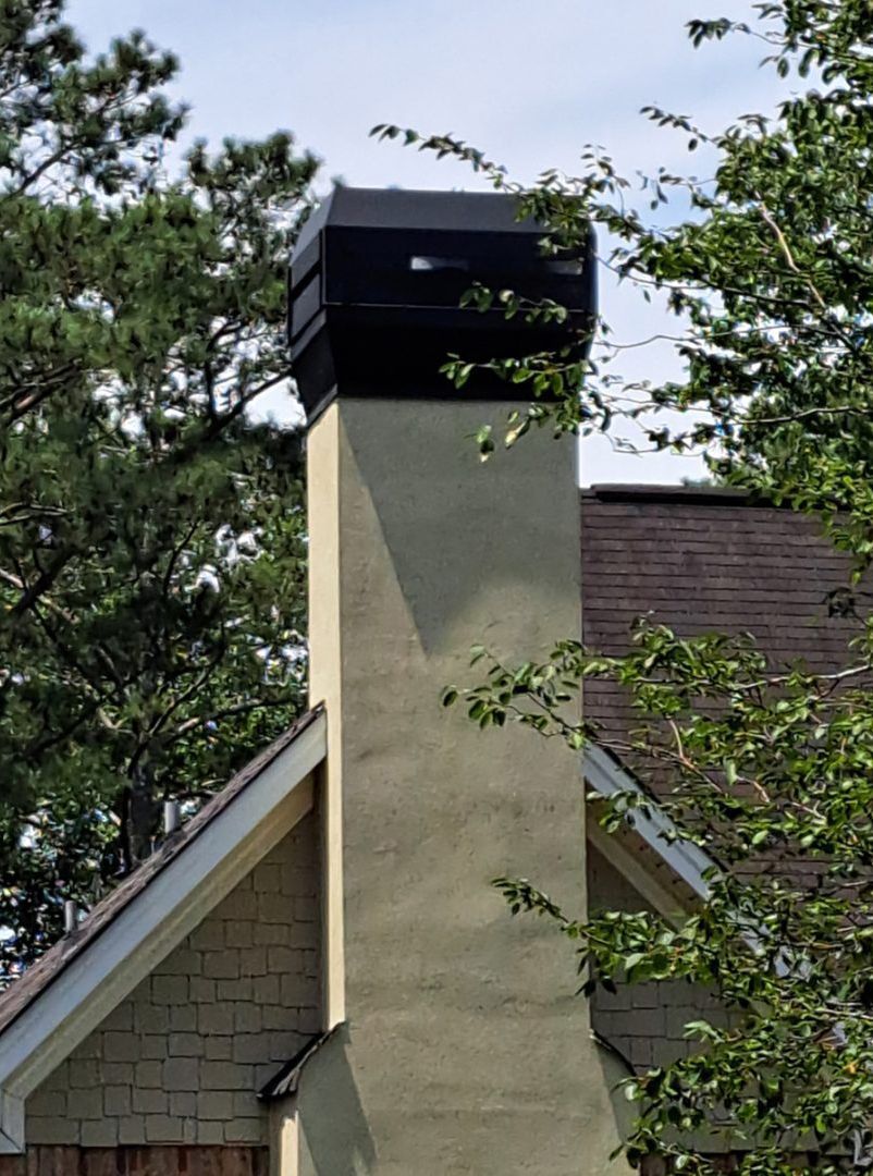 Tall, square chimney with a black cap on a house roof, framed by green trees.