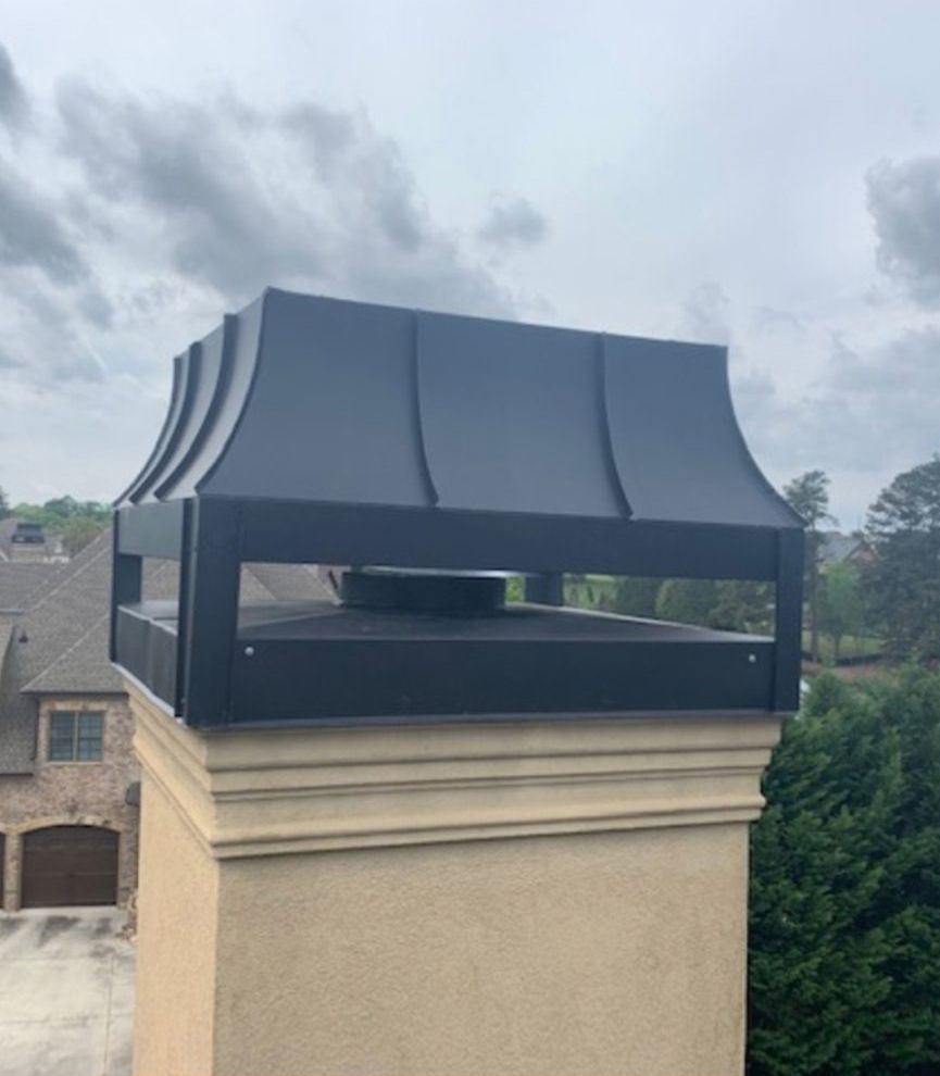 Black metal chimney cap on a beige stucco chimney with a cloudy sky background.