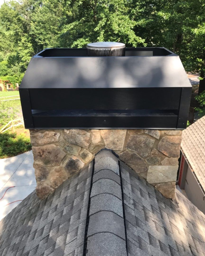 Black chimney cap atop a stone chimney on a shingled roof.