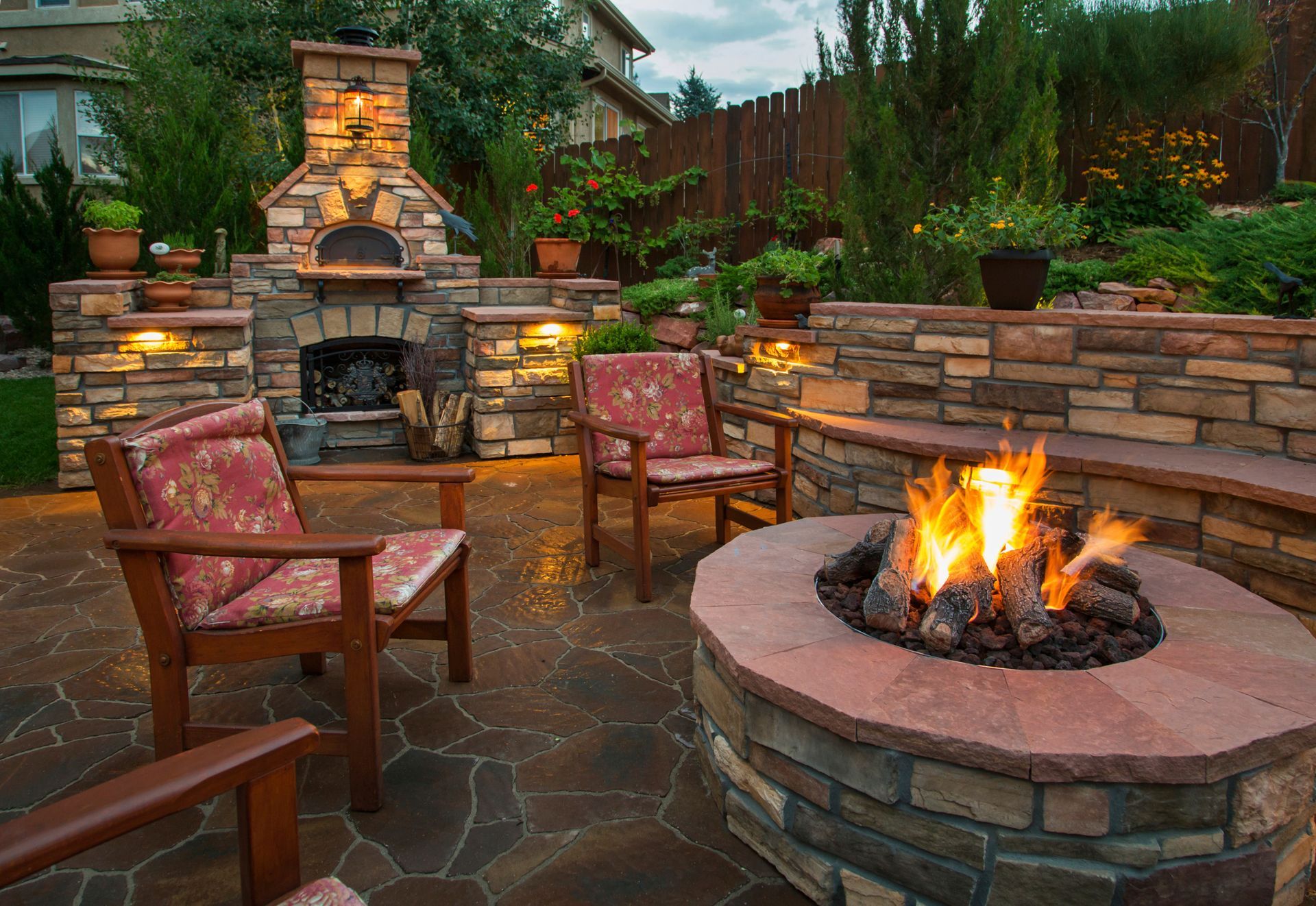 Backyard patio with lit fire pit and outdoor pizza oven, surrounded by stone features and seating.