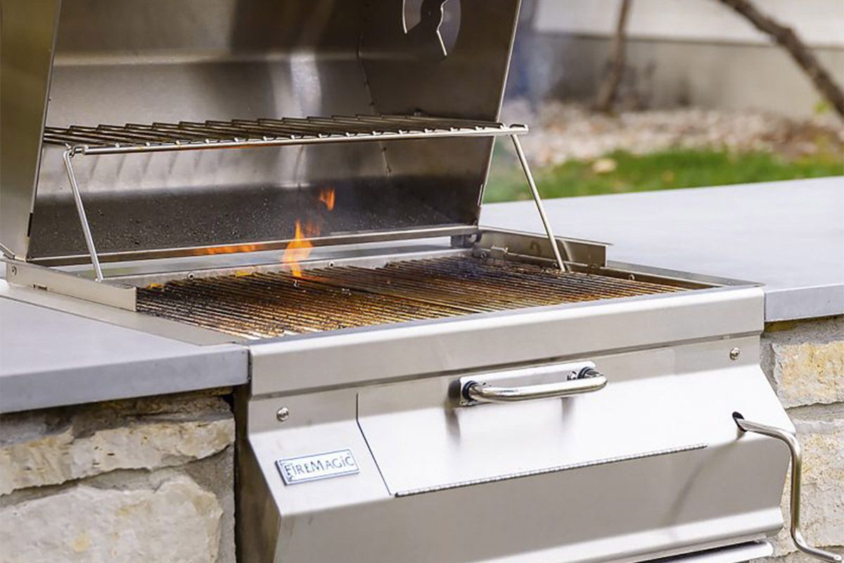 Outdoor stainless steel grill with flames rising from the grates, built into a stone and concrete surface.