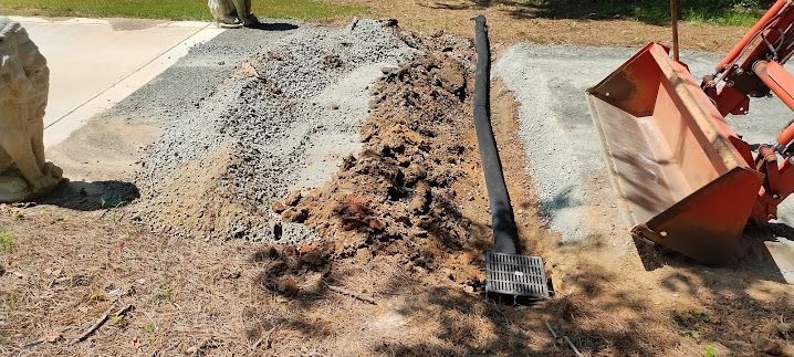 A trench being dug with a drainage pipe inside, next to a gravel path. An excavator is in the frame.