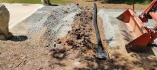 A trench being dug with a drainage pipe inside, next to a gravel path. An excavator is in the frame.