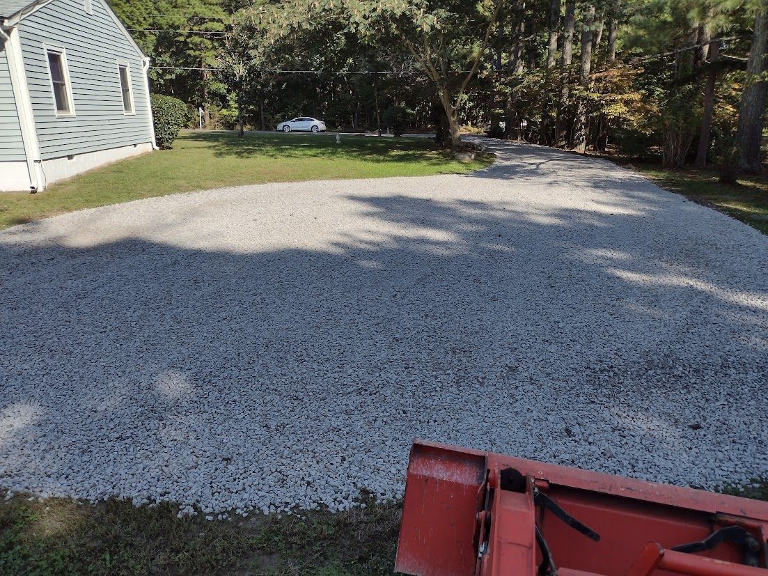 A gravel driveway in front of a house with an attached tractor bucket in the foreground.