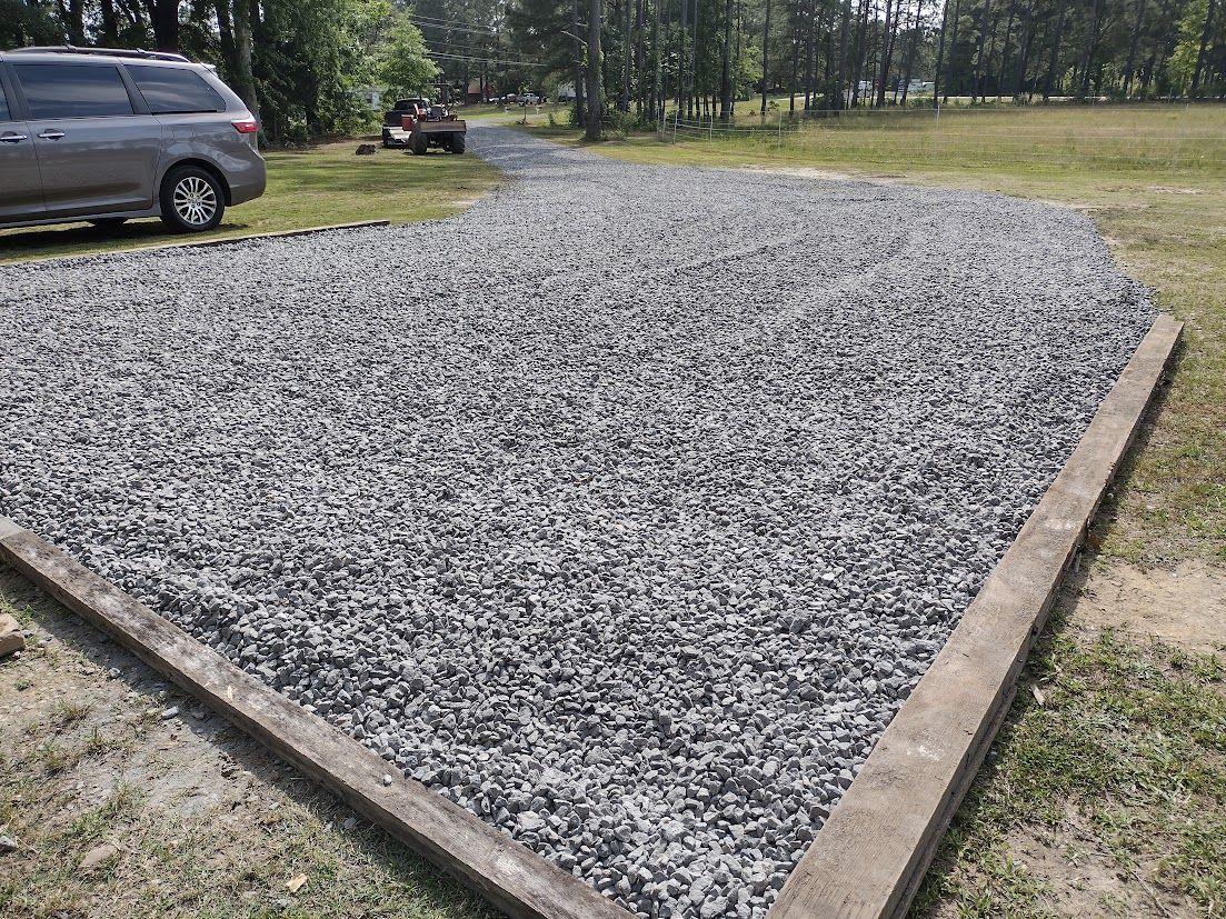 Gravel pad bordered by concrete, next to a car and grassy area. A vehicle is in the background.
