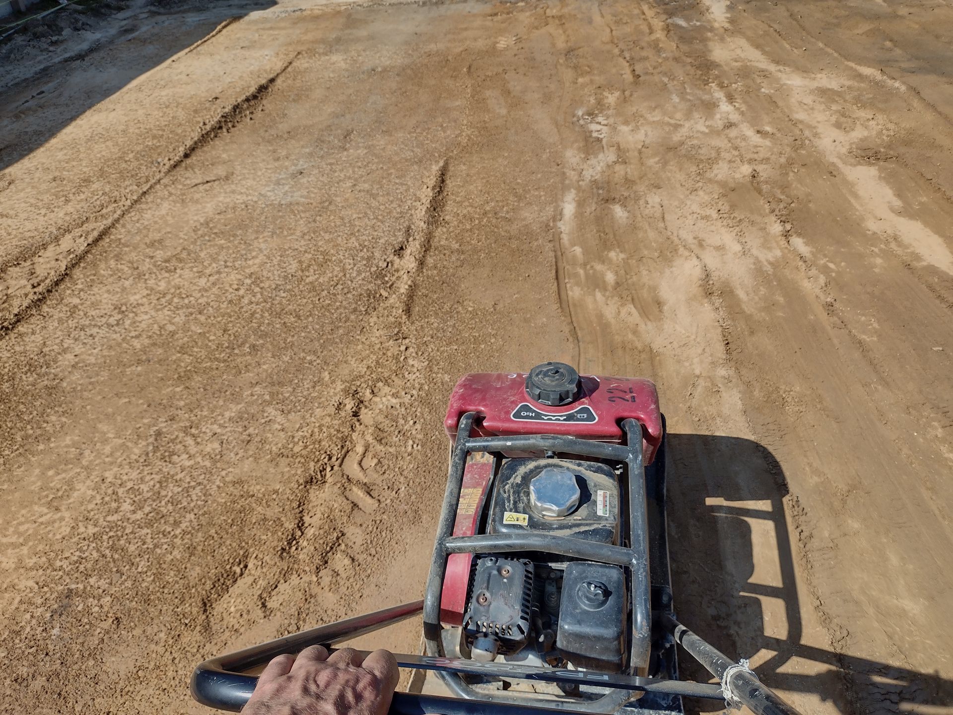 Man operating a red plate compactor on a sandy surface, compacting the ground.