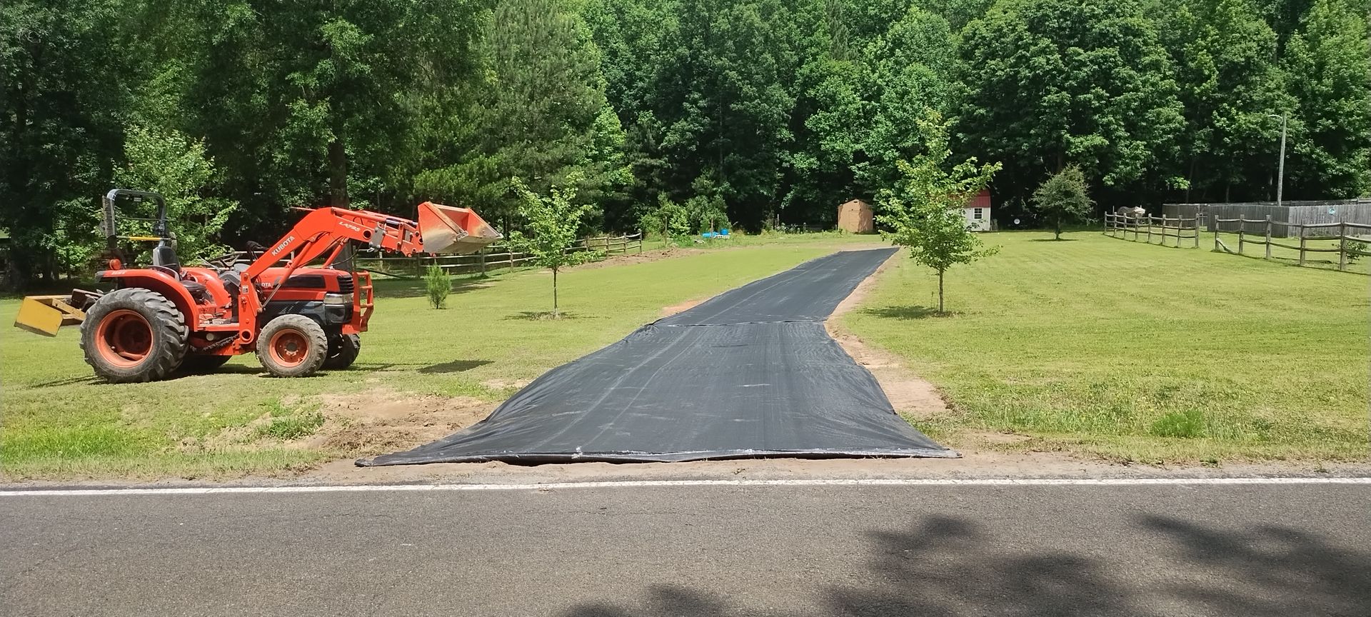 An orange tractor working on a black driveway, surrounded by trees and grass.