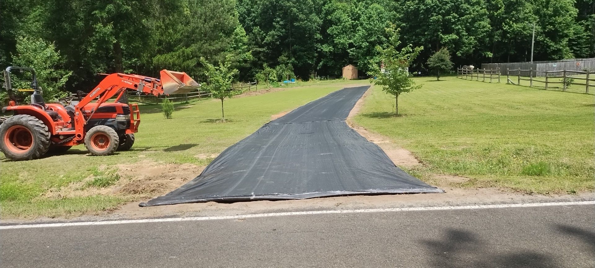 An orange tractor works on a driveway covered with black material, near a road, and green grass.
