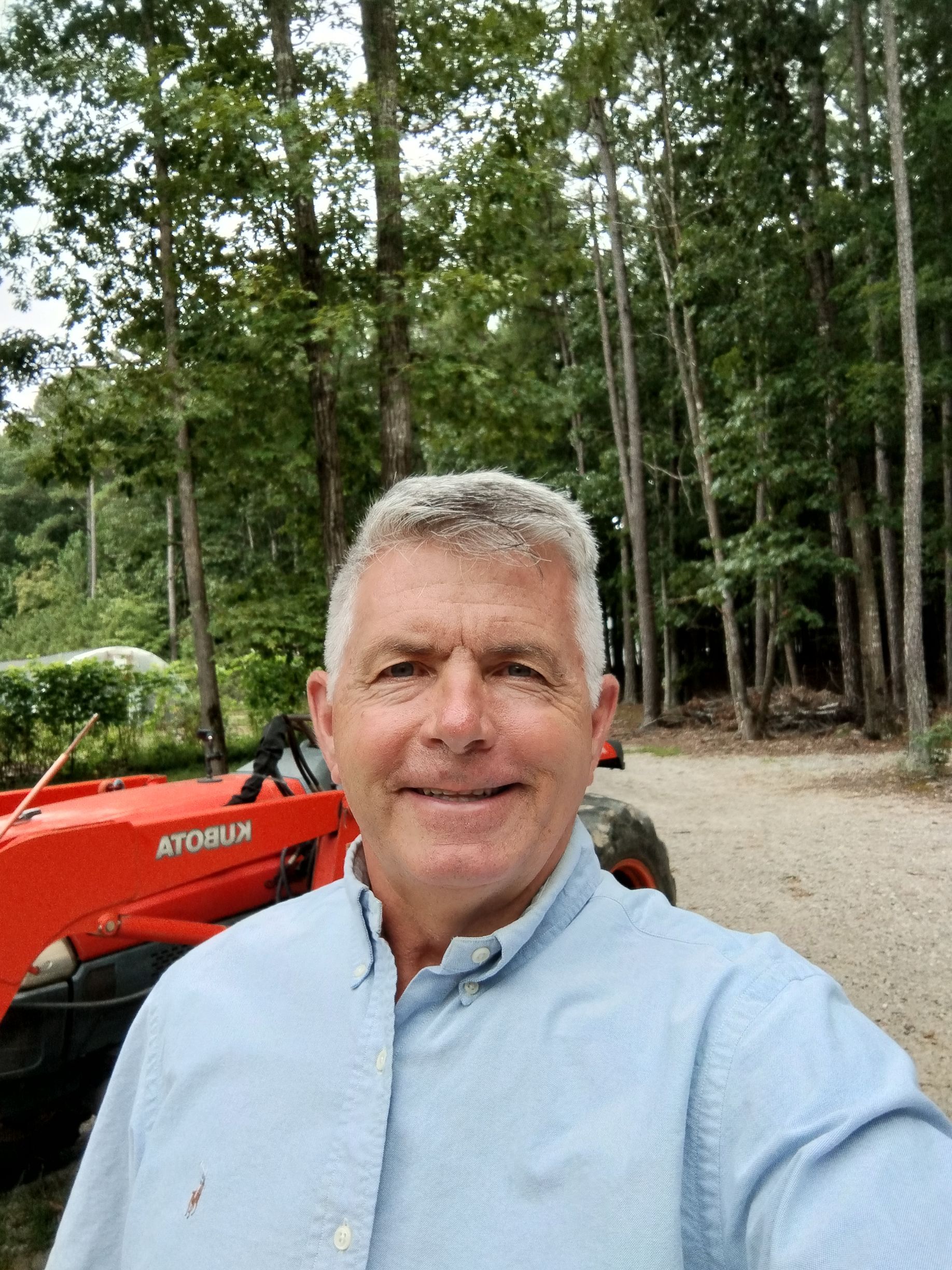 Man in light blue shirt smiles near red tractor in wooded area.