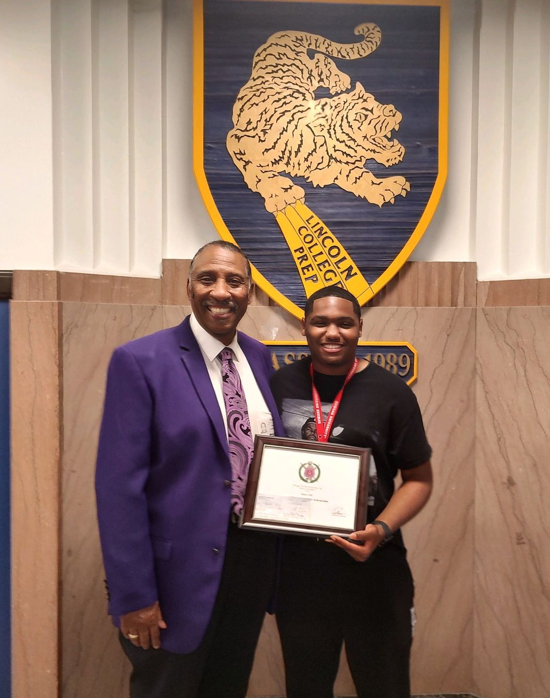 A man in a purple suit stands next to a young man holding a certificate