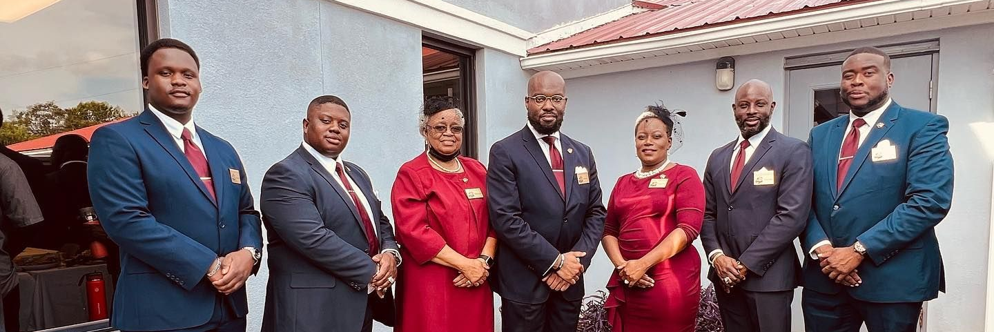 A group of seven people in formal business attire pose together outside a building with light blue walls.