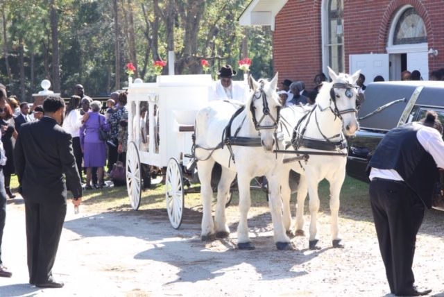 A white horse-drawn carriage hearse parked outside a brick building with people gathered nearby for a funeral service.