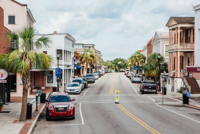 A street view in a historic town with cars parked along both sides, palm trees, and multi-story colonial-style buildings.