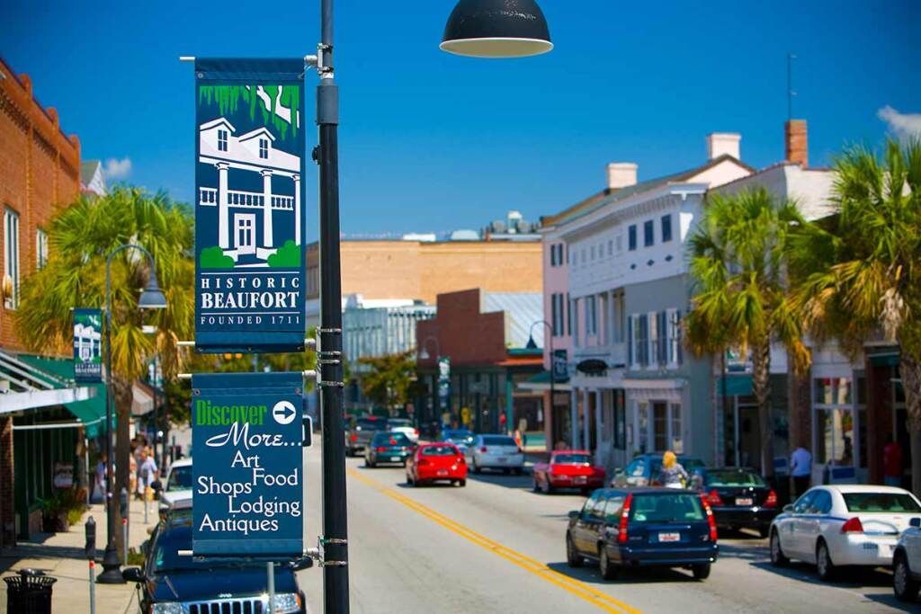 A street view of downtown Beaufort, South Carolina, with historic buildings, palm trees, and a banner on a light pole.
