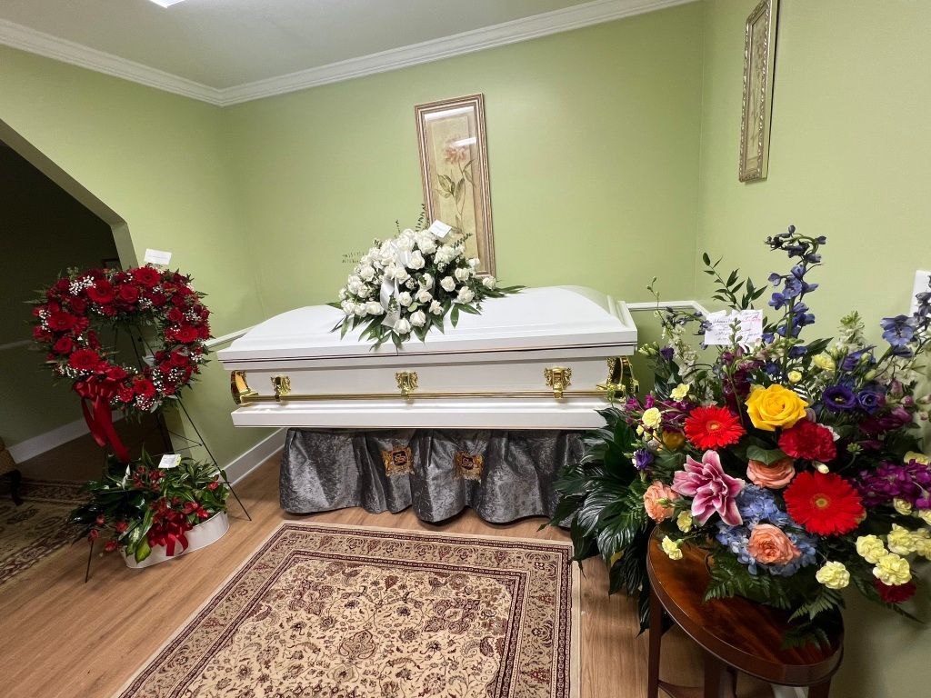A white casket centered in a room with a floral wreath to the left, a large bouquet to the right, and a patterned rug.