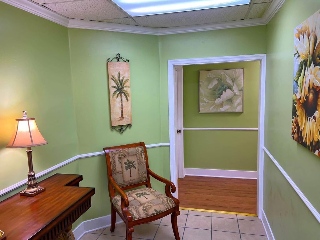 A hallway with light green walls, a wooden side table with a lamp, a palm-patterned chair, and framed wall art.