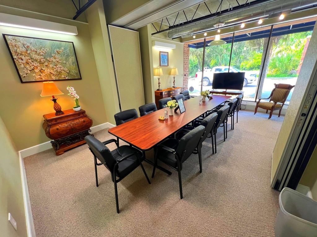 A conference room with a long wood-grain table, black chairs, patterned carpet, a side table with a lamp, and a window view.