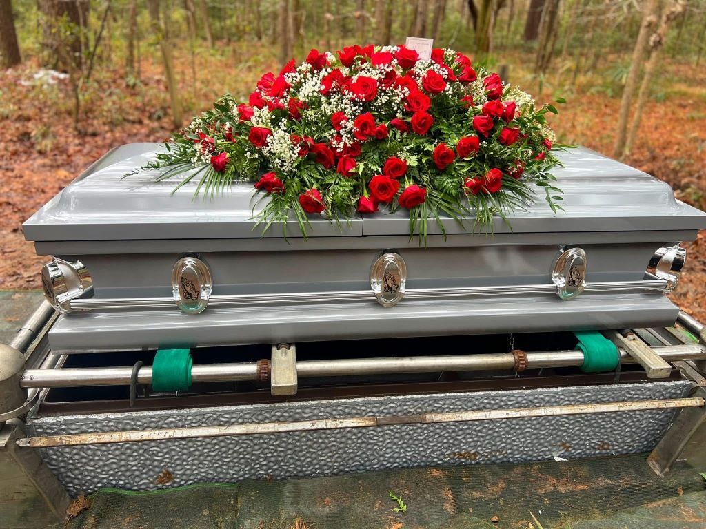 A grey metal casket topped with a vibrant arrangement of red roses and white filler flowers, resting on a grave marker.