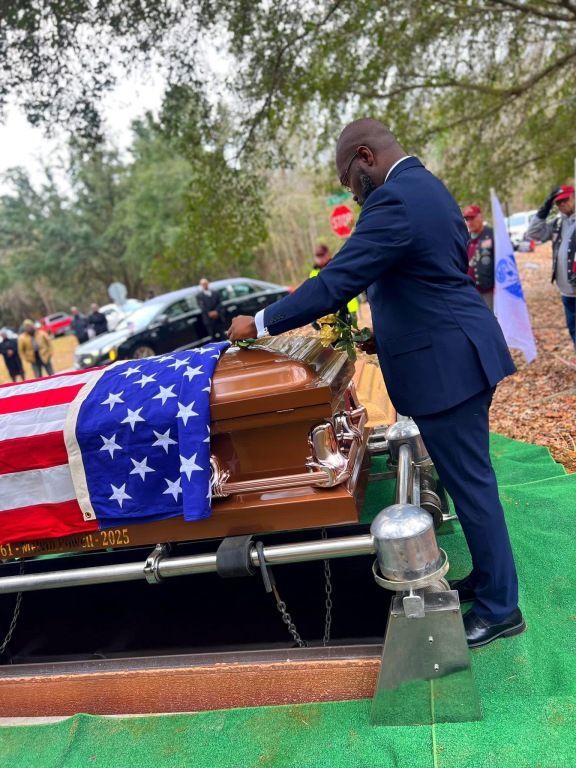 A person in a dark suit places a hand on a casket draped in an American flag during an outdoor graveside service.