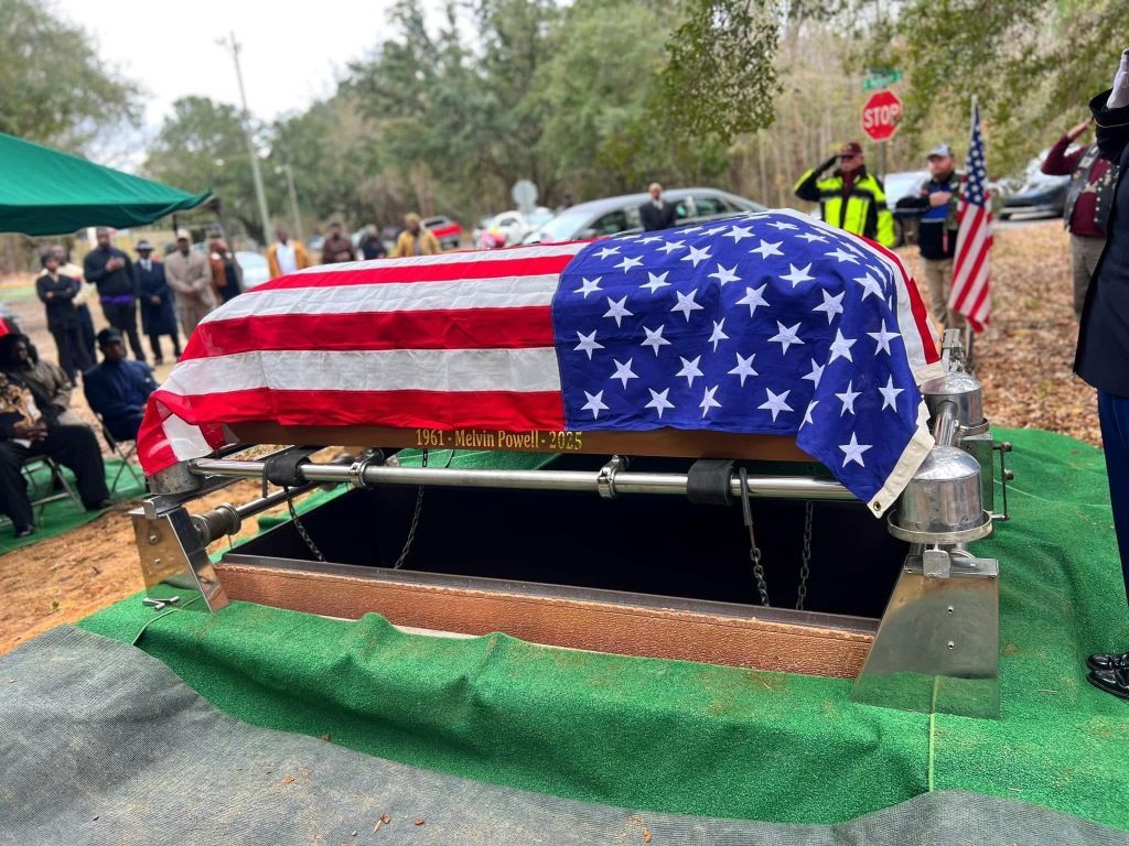 A casket draped in an American flag sits over an open grave at a burial service, with mourners standing nearby.
