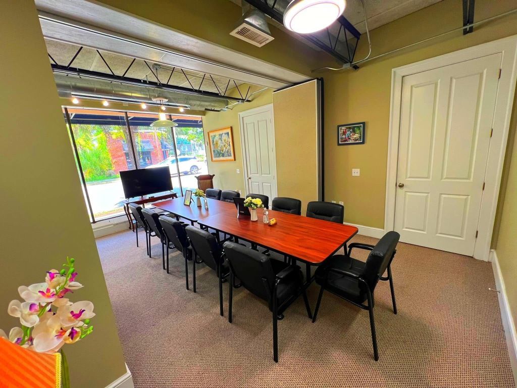Conference room with a long wood table and black chairs, large windows, tan walls, and a white door.