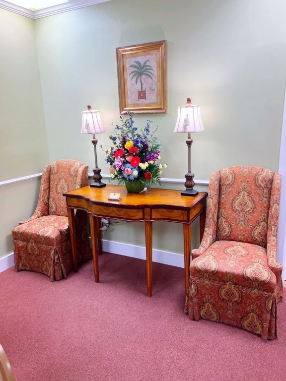 Two patterned upholstered chairs flank a wooden side table with two lamps and a colorful floral centerpiece.