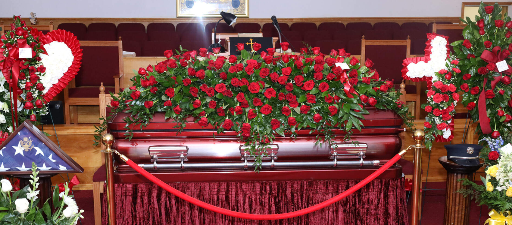 A casket draped in a large arrangement of red roses sits at the front of a sanctuary, flanked by floral displays and a flag.