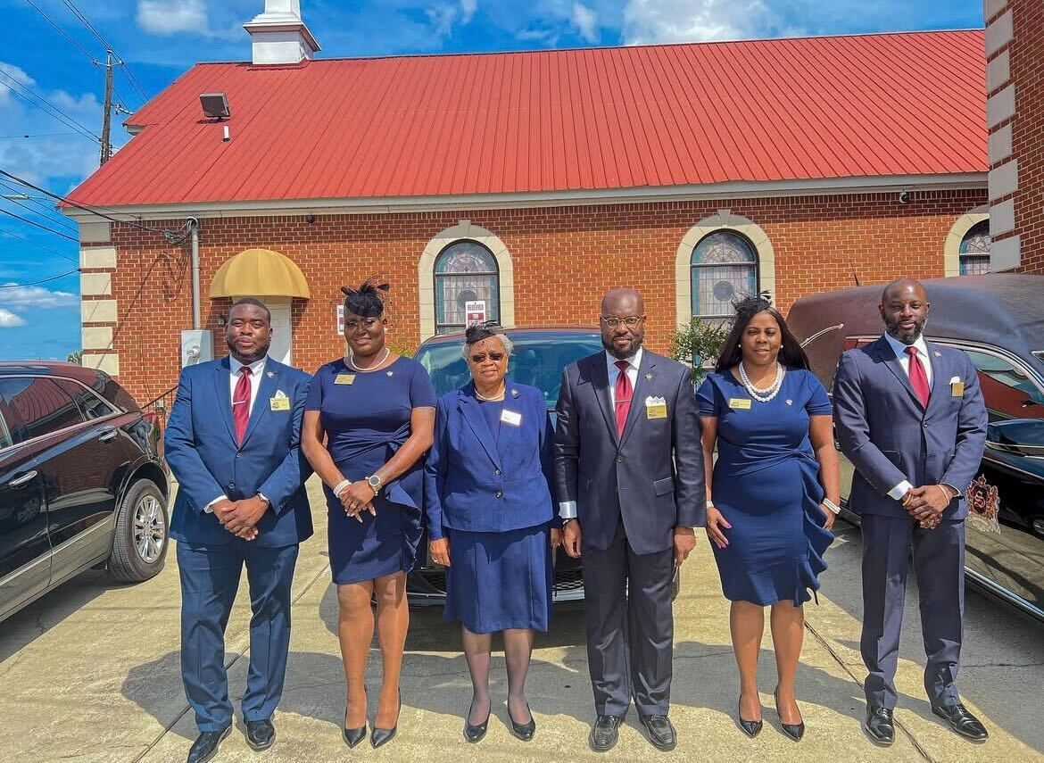 Five professionals in formal dark suits stand in a row outdoors in front of a red-roofed brick building.