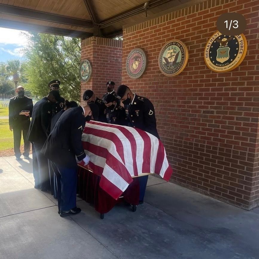 Military honor guard members in uniform drape an American flag over a casket at an outdoor memorial site.