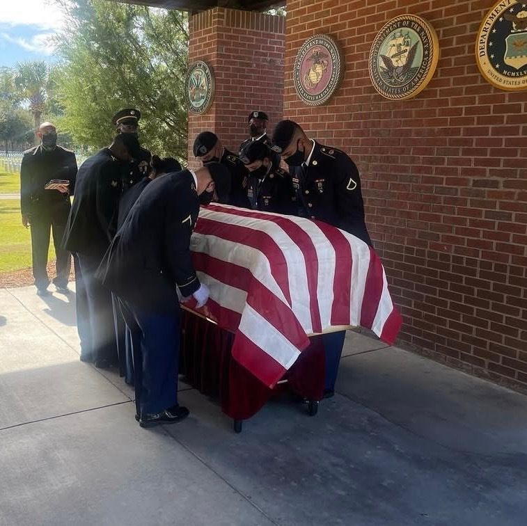 Military personnel in dress uniforms perform a ceremonial flag-draping of a casket at a brick memorial site.
