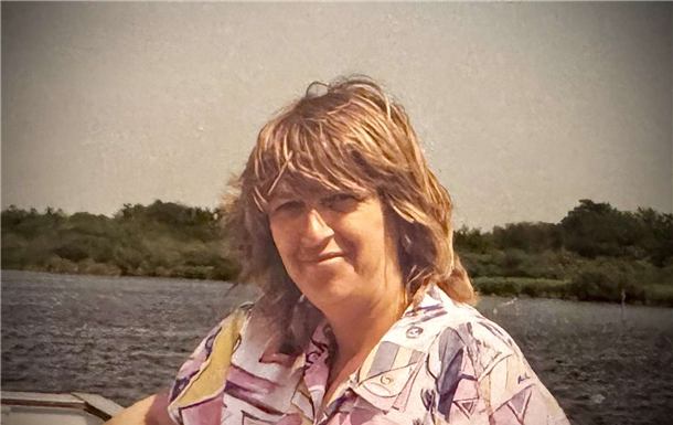 Woman with light brown hair in patterned shirt near a body of water and treeline.