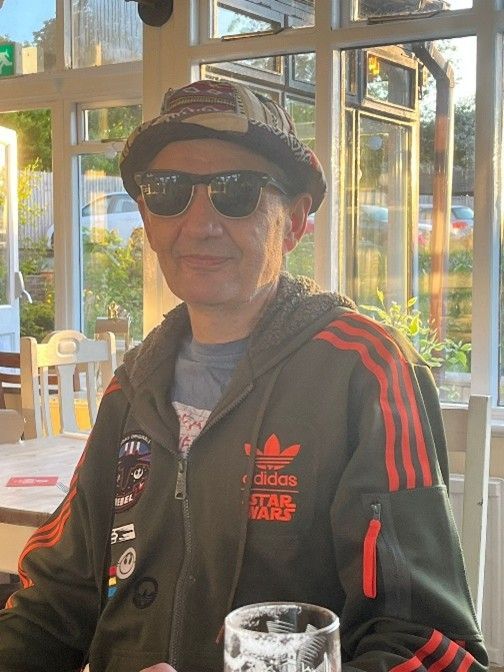 Man wearing sunglasses, hat, and Star Wars Adidas jacket, sitting inside at a table near a window.