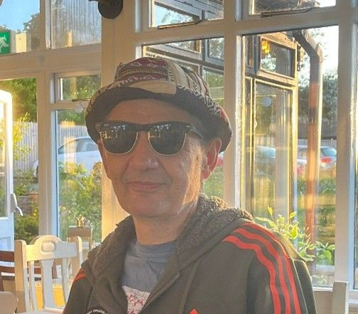 Man wearing sunglasses and hat, in an Adidas Star Wars jacket, sitting at a table near a window.