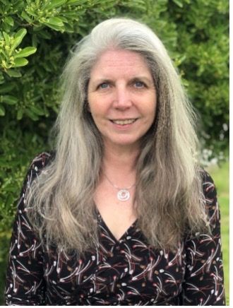 Woman with long, gray hair, smiling, wearing a patterned top, outdoors near green foliage.
