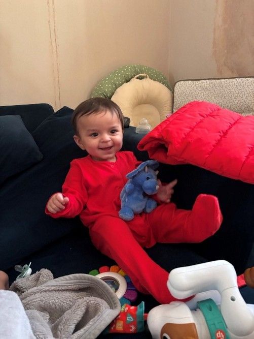 Smiling child in red pyjamas sitting on a couch, holding a blue toy, surrounded by pillows and toys