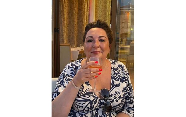 Woman holding a glass of wine, smiling, wearing a blue and white patterned dress, seated indoors.