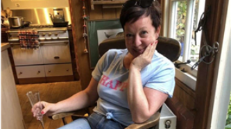 Woman in blue shirt, sitting in chair with hand on chin; kitchen in background.