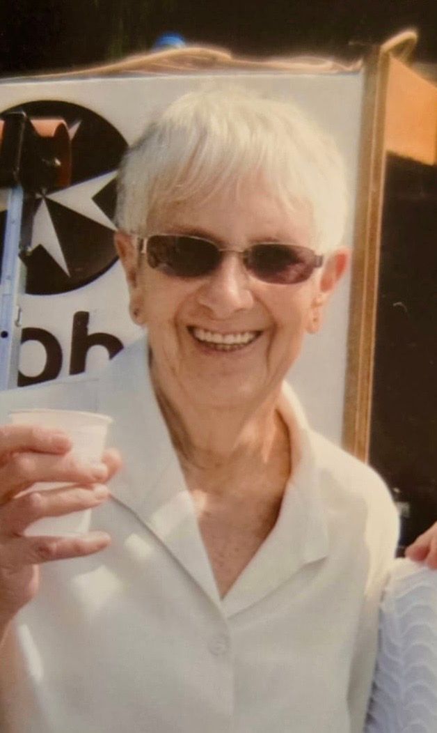 Woman with short white hair and sunglasses smiles, holding a cup outside.