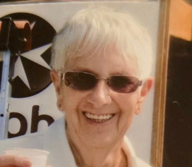 Woman with short white hair, wearing sunglasses and white shirt, smiles, holding a cup outside.