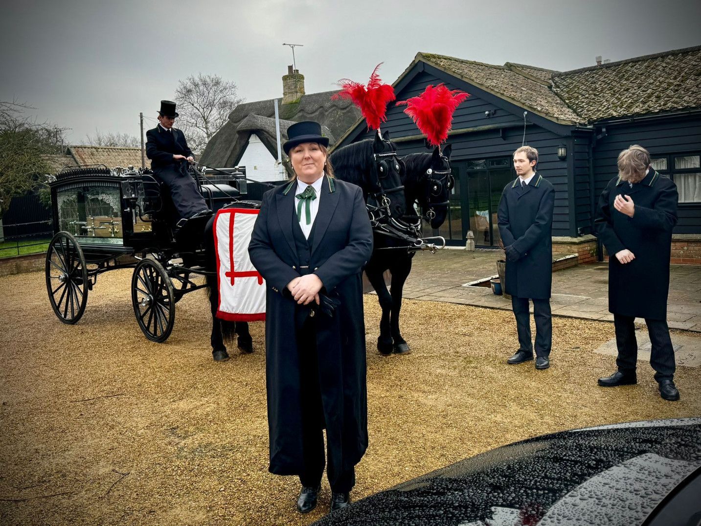 A woman in a top hat stands in front of a horse drawn carriage