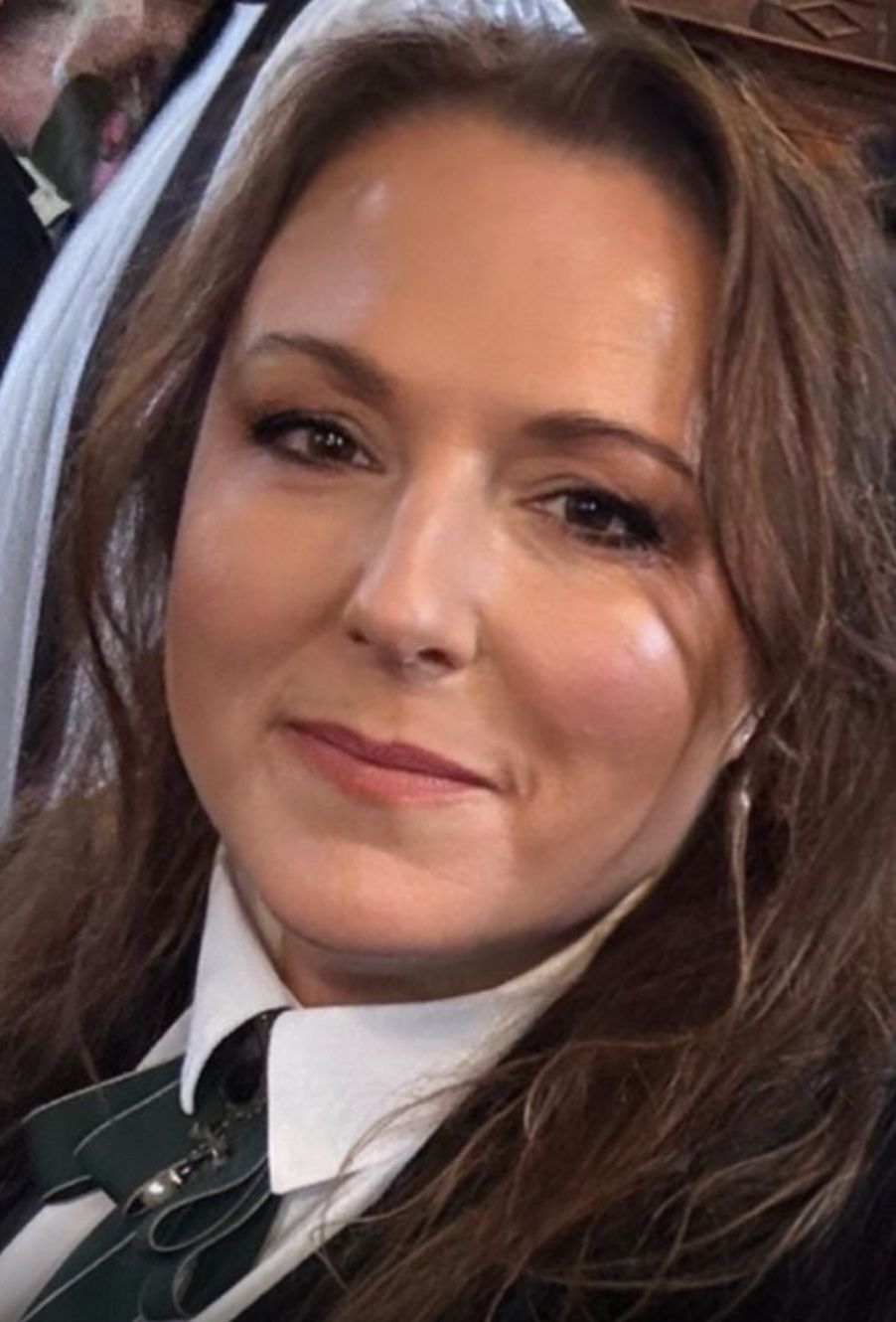 Woman with brown hair smiles, wearing a white collared shirt with a green tie, indoors.
