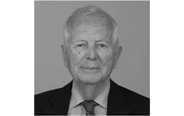 Studio headshot of an older man in a suit and tie against a plain gray background