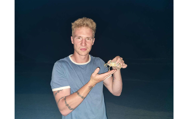 Man with blonde hair holding a crab; wearing blue shirt with white trim; against a dark blue background.
