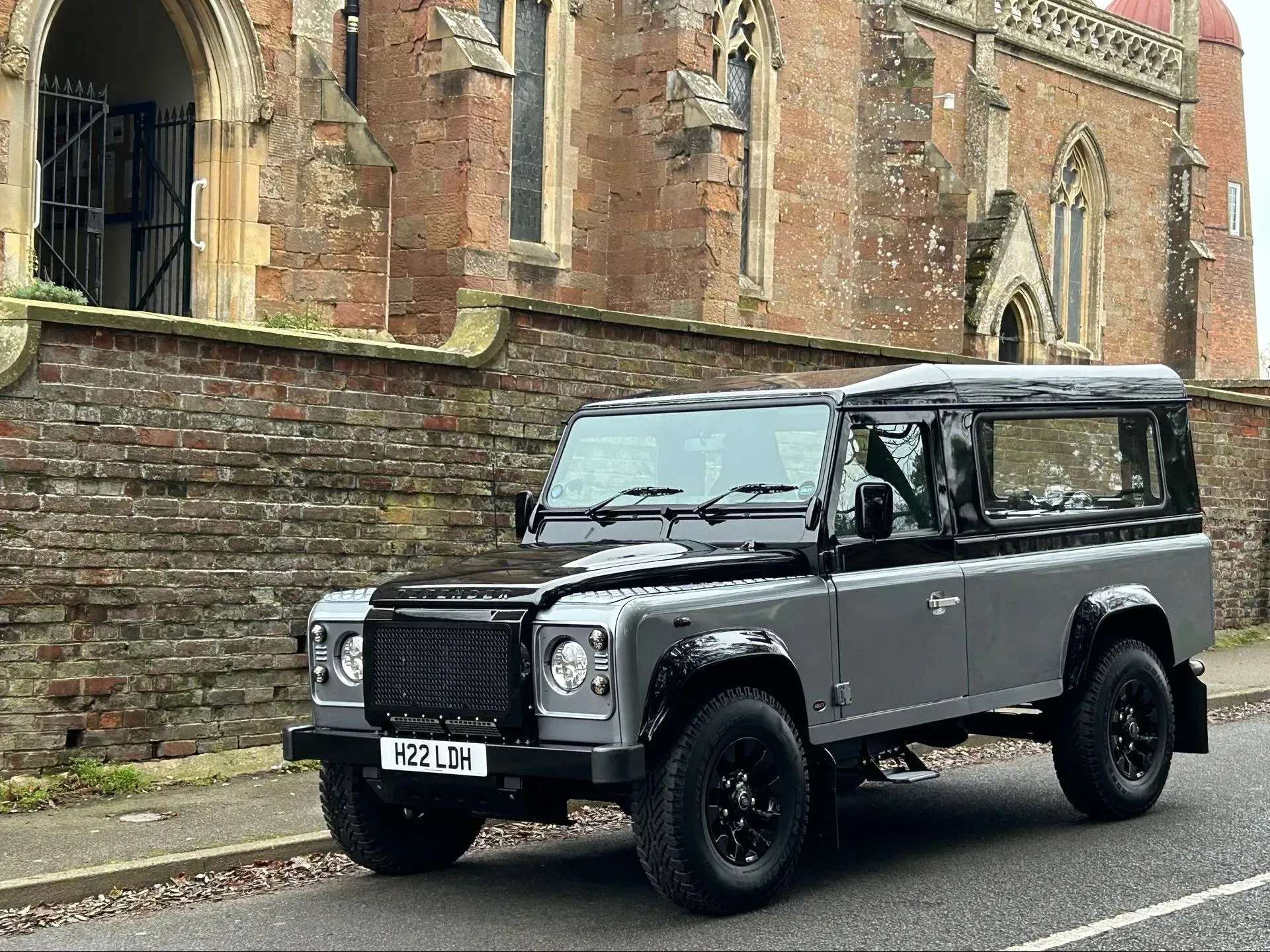 A gray land rover defender is parked on the side of the road in front of a brick building.