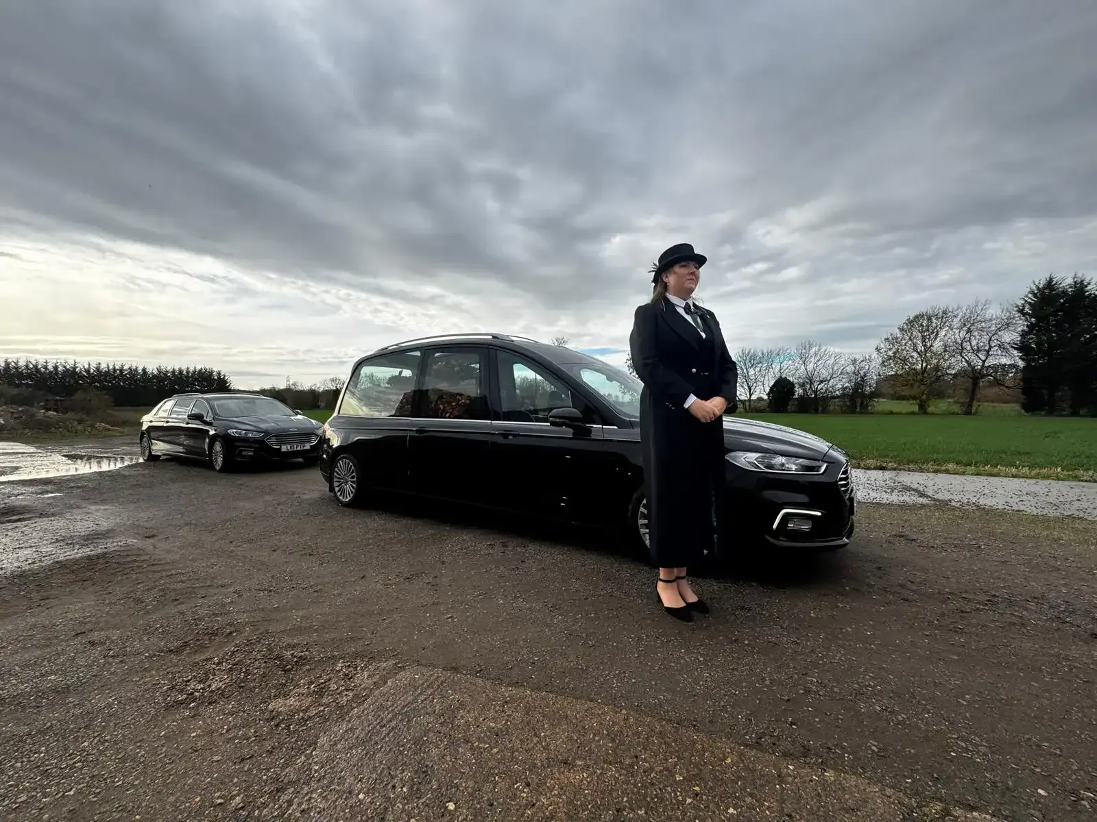 Woman in black suit stands by black car on gravel road under cloudy sky, another car in background.