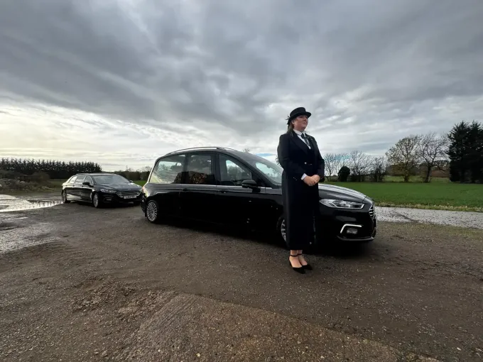 Woman in black suit and hat standing by black car on a cloudy day. Another black car in background.