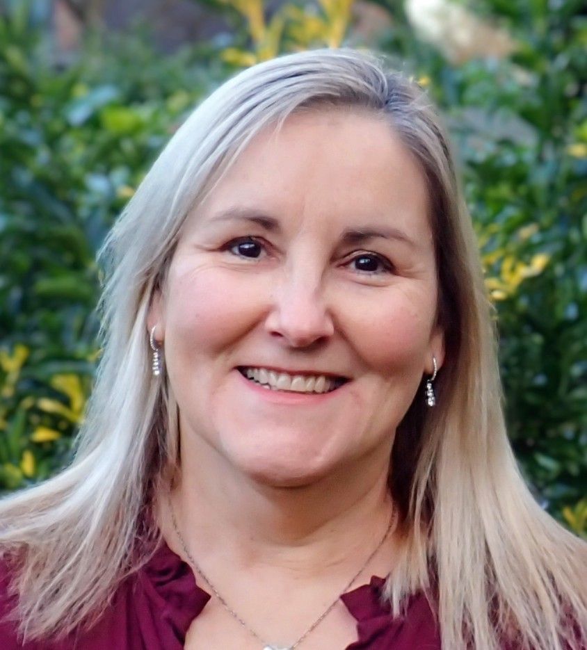 Woman with long blonde hair, smiling, wearing a dark red blouse, outdoors with green foliage in background.